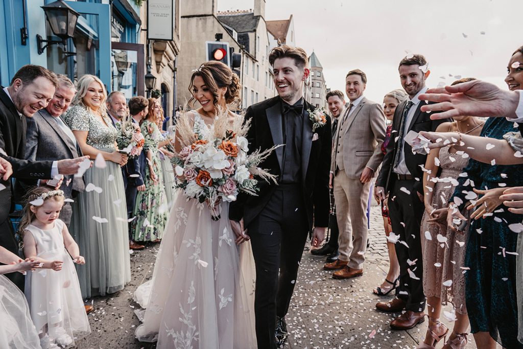 Exterior shot of bride and groom on their wedding day outside the granary with guests throwing petals and watching them walk towards the camera